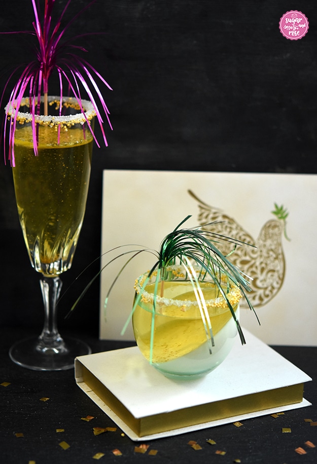 Ein hohes Sektglas mit Sektjelly und pink Deko, daneben auf einem weißen Buch stehend ein kleines bauchiges Glas mit Sektjelly und grüner Deko, dahinter eine Karte mit Friedenstaube als Motiv