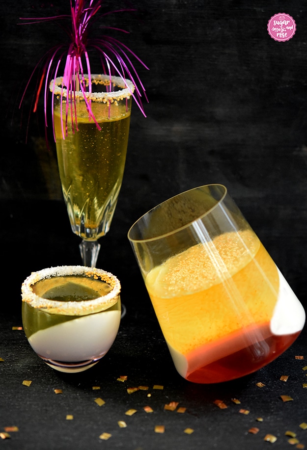 Ein Sektglas mit Sektjelly und pinkfarbener Cocktaildrink-Deko, davor ein bauchiges Glas mit Sektjelly und Wodka-Pannacotta, daneben ein Glas mit dreifarbigem Sektjelly (gold, weiß, rot)