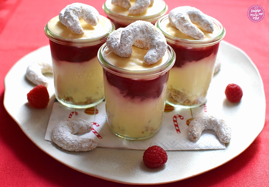 Vier Gläser mit Vanillekipferlmousse auf einem großen Keramikteller, dekoriert mit Vanillekipferln und Himbeeren und Weihnachtsserviette auf einem knallroten Weihnachtsgeschirrtuch stehend – in Großaufnahme, sodass die Schichten zu sehen sind: unten Vanillekipferl, darauf helles Mousse, dann eine rote Fruchtschicht, darüber wieder Mousse und obenauf jeweils zwei Vanillekipferl als Dekoration