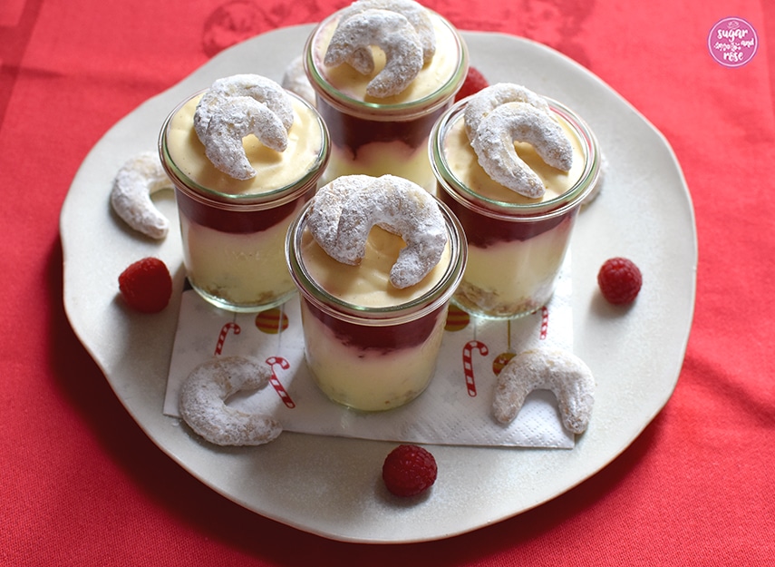 Vier Gläser mit Vanillekipferlmousse auf einem großen Keramikteller, dekoriert mit Vanillekipferln und Himbeeren und Weihnachtsserviette auf einem knallroten Weihnachtsgeschirrtuch stehend