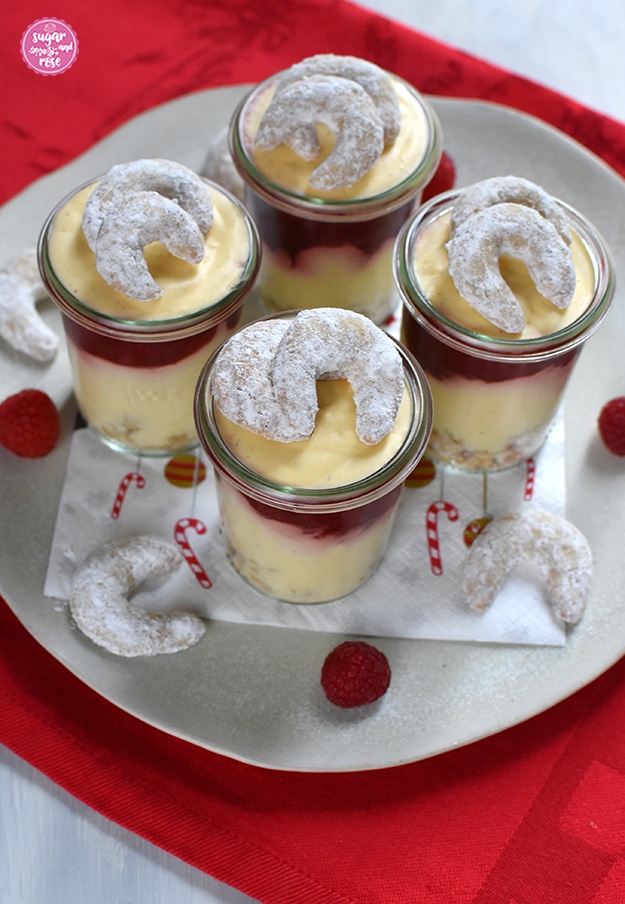 Vier Gläser mit Vanillekipferlmousse auf einem großen Keramikteller, dekoriert mit Vanillekipferln und Himbeeren, auf einem knallroten Weihnachtsgeschirrtuch stehend