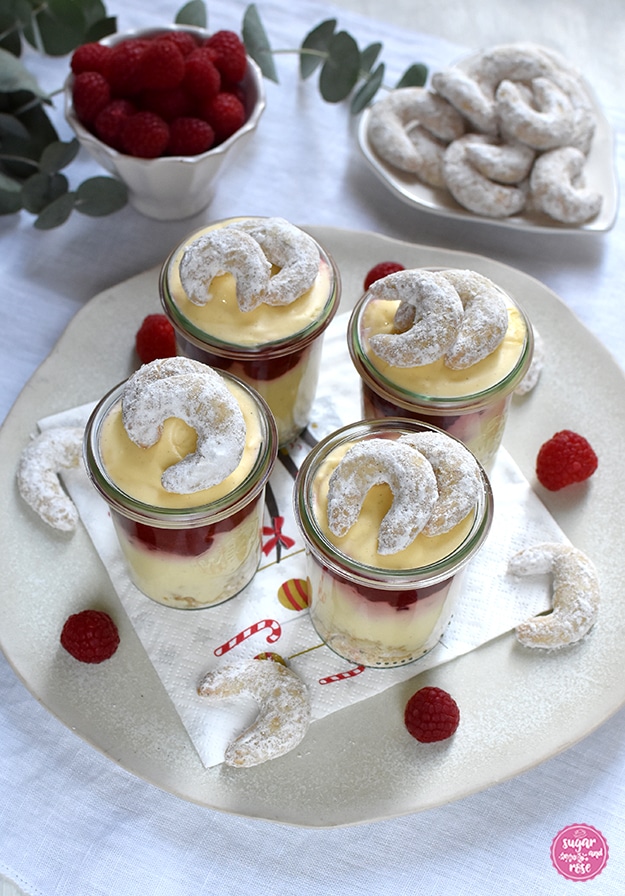 Vier Gläser mit Vanillekipferlmousse auf einem großen Keramikteller, dekoriert mit Vanillekipferln und Himbeeren, dahinter eine Schale frischer Himbeeren und ein herzförmiger Teller mit Vanillekipferln