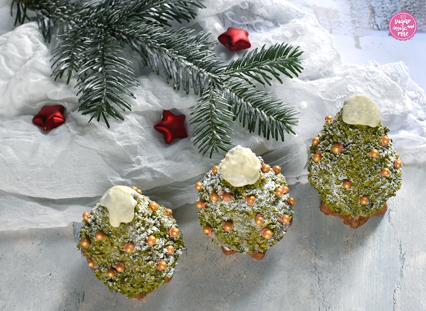 Drei Weihnachtsbäume zum Vernaschen mit Goldzuckerperlendeko in Reih und Glied vor einem weißen Spitzentuch und einem beschneiten Tannenzweig, dazwischen drei mattrote Dekosterne