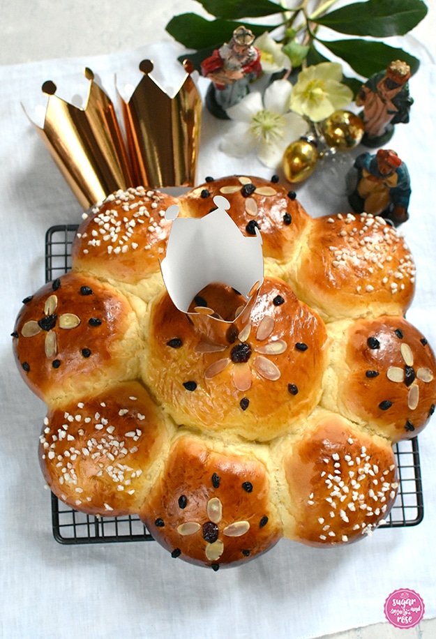 Dreikönigskuchen auf Gitterrost mit einem Goldkrönchen, dahinter zwei weitere Krönchen, Schneerosenblüten und die drei Königsfiguren