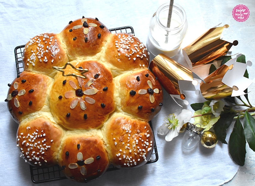 Fertig gebackener Dreikönigskuchen, in der Mitte ein sternförmiger Sternspritzer aufgesteckt, rechts daneben drei Goldkrönchen liegend, zwei Goldkugeln und eine Glaskugel, im Hintergrund ein Milchglas