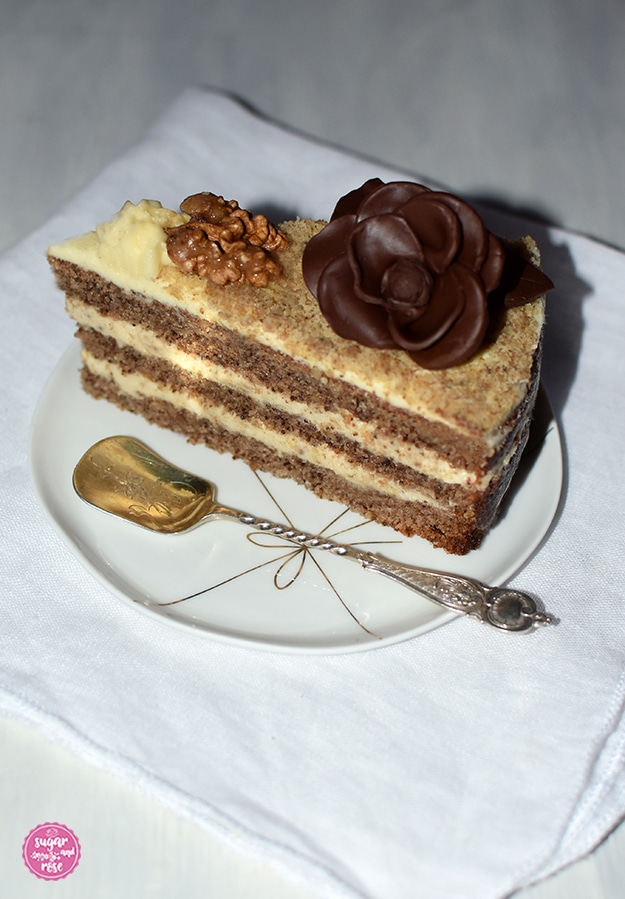 Ein Stück Walnusstorte auf einem kleinen weißen Porzellanteller mit goldener Masche als Dekor, dazu ein alter englischer Silberlöffel. Das Tortenstück mit karamellisierter Walnusshälfte und einer Schokoladenrosenblüte wird auf einer weißen Leinenserviette präsentiert.