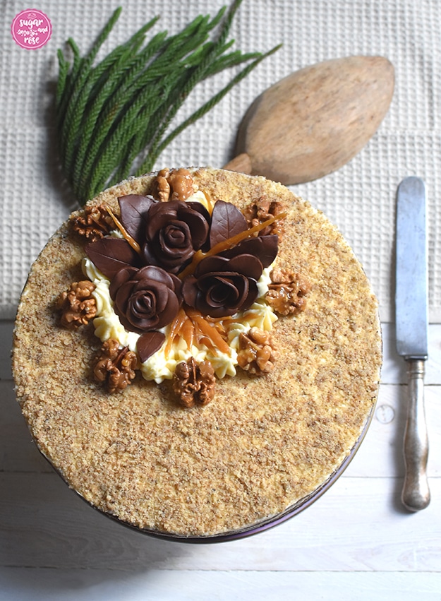 Walnusstorte mit karamellisierten Walnüssen, Schokoladerosen von oben auf einem naturfarbenen Waffelpiquetuch, daneben ein altes Messer, als Deko ein grüner Zweig und eine alte hölzerne Mehlschaufel