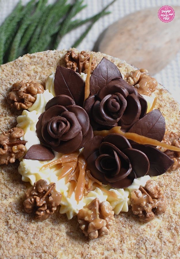 Detailausschnitt der Dekoration auf der Walnusstorte: ein weißes Cremebett auf einem Teil der Oberfläche, darauf drei Schokoladenblüten mit Blättern, dazwischen Krokantstangen, rund um die Creme die karamellisierten Walnusshälften