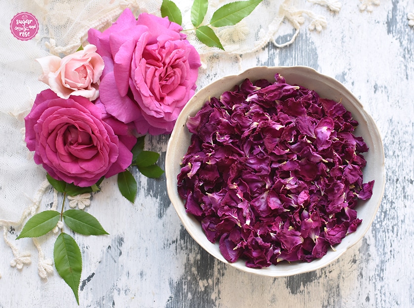 Eine Schale mit getrockneten dunkelrosa Blüten der Rose de Resht, dahinter drei frische rosa Rosenblüten, grüne Rosenblätter und ein zartes cremefarbenes Vintagetuch
