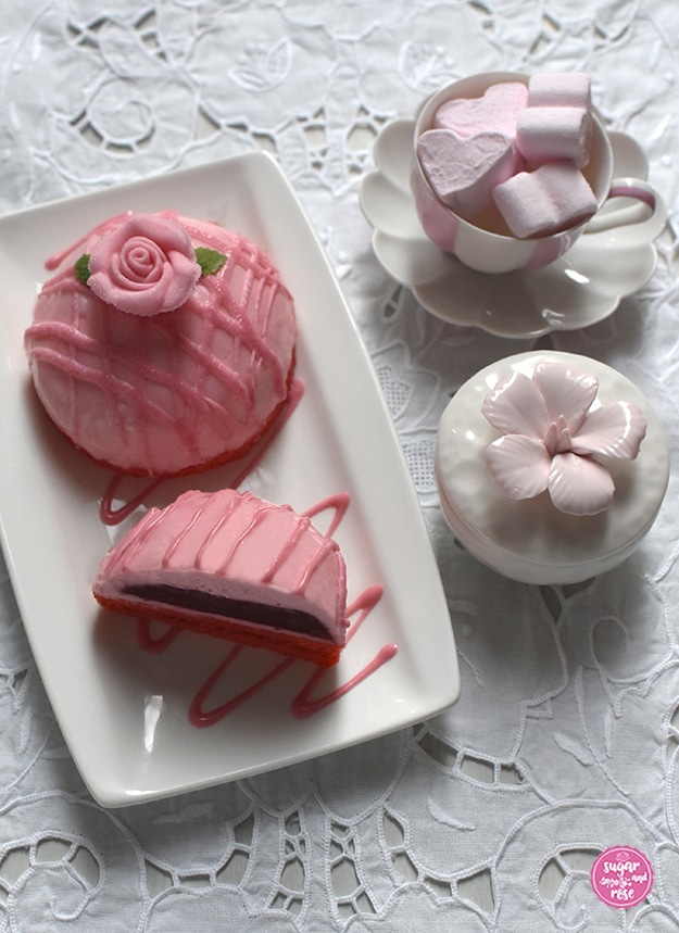 Ein Stück Himbeer-Joghurt-Törtchen neben einem halben Stück auf einem rechteckigen weißen Teller. Daneben eine Augarten-Melone-Mokkatasse mit rosa Marshmallowherzen und eine kleine weiße Keramikschale mit Blütendeckel.