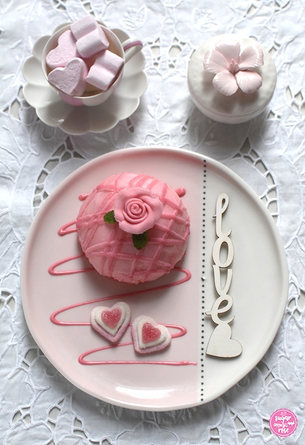 Ein Himbeer-Joghurt-Törtchen mit Dekorose auf rosa-weißem Keramikteller, dekoriert mit einem Love-Schriftzug, dahinter eine Augarten-Melone-Mokkatasse mit rosa Marshmallowherzen