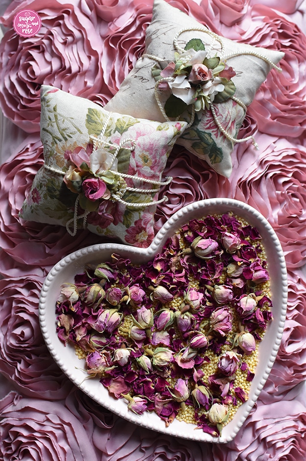Rosa Herzschale mit Hirsespelzen und Rosenknospen, dahinter auf einem Kissen mit Seidenrosen als Oberfläche zwei quadratische Rosenduftkissen mit Kordeln und Seidenblütendekoration