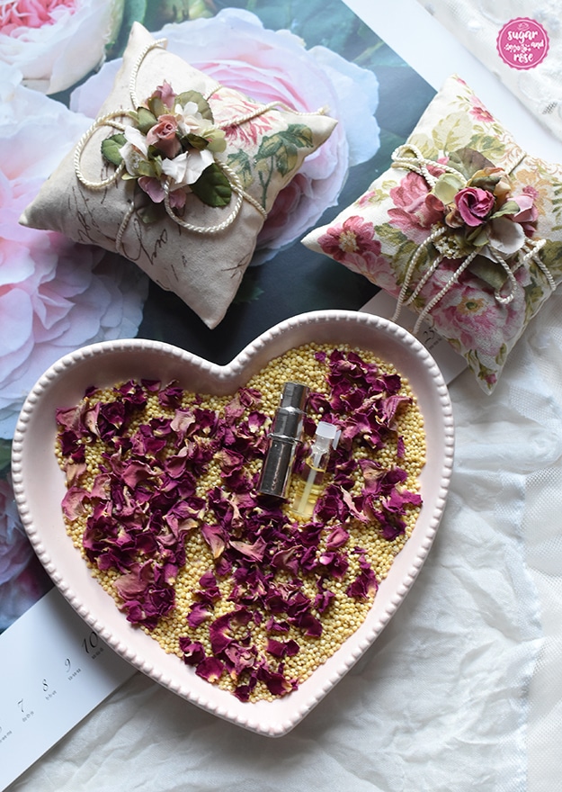 Zwei quadratische Rosenkissen aus Rosendekorstoff, umwickelt mit weißer Kordel, dekoriert mit Seidenblüten, davor eine rosa herzförmige Schale mit Hirsespelzen, getrockneten Rosenblüten und Rosenöl-Phiolen