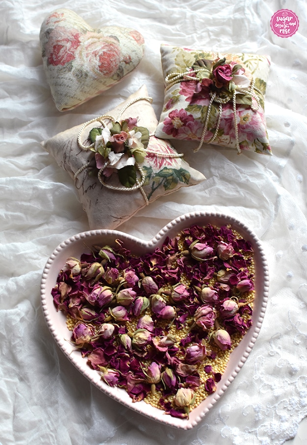 Im Vordergrund eine dezent rosa Herzschale mit Hirsespelzen und Rosenknospen, dahinter auf einem weißen Spitzenschal zwei quadratische und ein herzförmiges Rosenduftkissen mit Rosendekorstoff