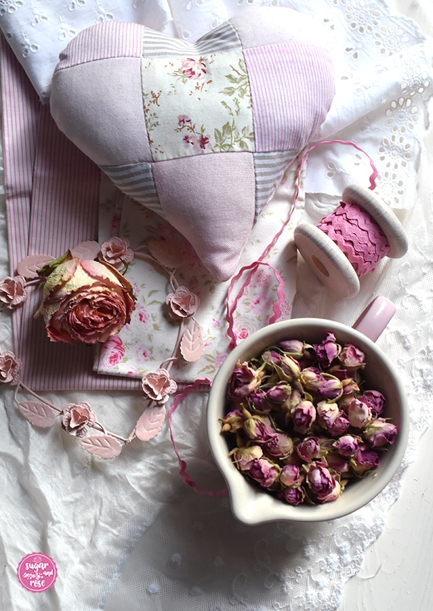 Rosenduftkissen in Herzform aus rosa-weiß gestreiftem, rosafarbenen und geblümten Stoffresten im Patchworkstil, davor eine Schale mit getrockneten Rosenblüten, Stoffreste und ein rosa Stoffband auf Spule