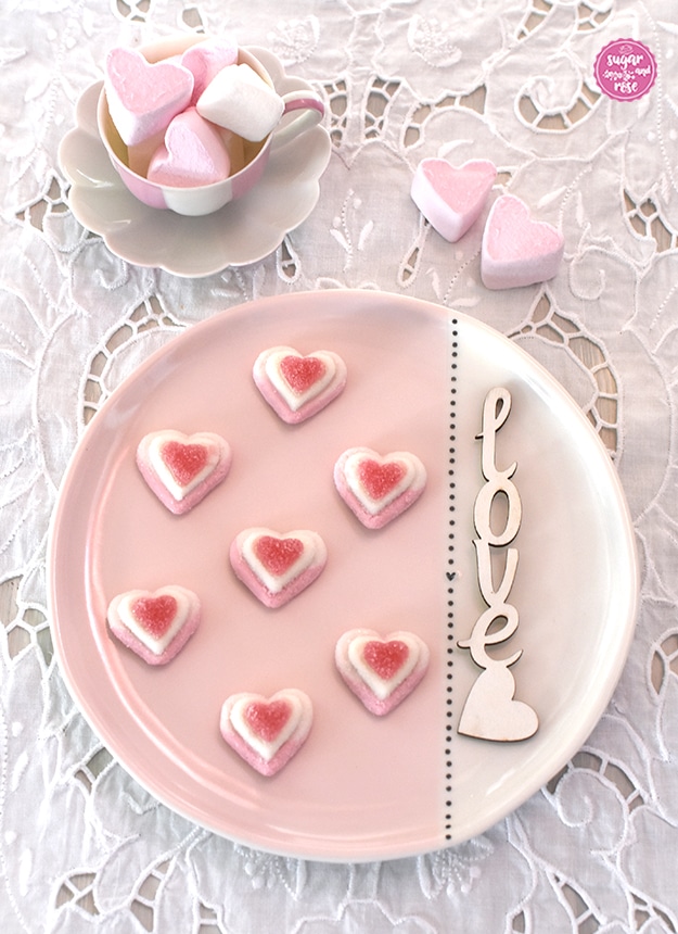 Rosa-weißer Keramikteller mit sieben dekorativ aufgelegten Zuckerherzen, dahinter eine Augarten-Melone-Mokkatasse mit rosa Marshmallowherzen gefüllt auf weißem Lochstickerei-Tischset