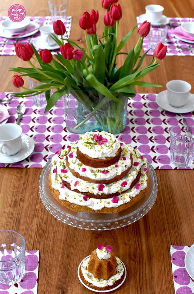 Frühstückstisch mit sechs Gedecken in weißem Porzellan, in der Mitte eine grüne Glasvase mit roten weinroten Tulpen, davor der Karottenkuchen mit Frischkäsefrosting auf der Glasetagere und davor ein kleines Küchlein