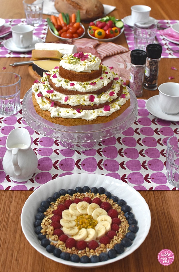 Frühstückstisch (Holz) mit rosa-weinroten nordischen Tischsets, in der Mitte die Glasetagere mit dem Karottenkuchen mit Frischkäsefrosting, davor eine weiße Schale mit Birchermüsli, dekoriert mit Heidelbeeren, Himbeeren und Banane. Im Hintergrund eine große runde Platte mit Schinken und Käse, Teller mit buntem Gemüse und ein großes Bauernbrot