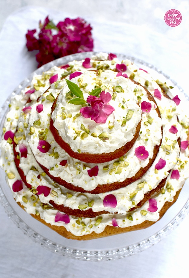 Karottenkuchen mit Frischkäsefrosting auf Glasetagere, dahinter eine pinke Rosenblütendolde
