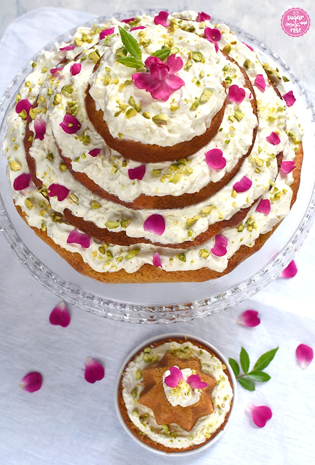 Karottenkuchen mit Frischkäsefrosting auf Glasetagere von vorne, davor ein kleines Küchlein auf einem extra Teller, rundum pinke Rosenblüten verstreut 