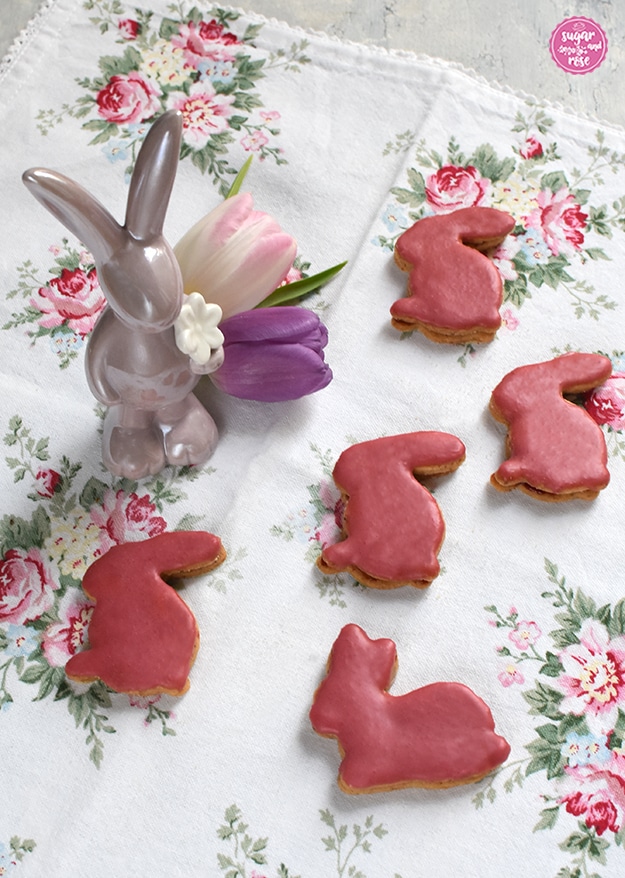 5 Osterhasen-Kekse in dunkelrosa glasiert auf einem Geschirrtuch mit Rosenblüten, dabei ein fliederfarbener Porzeallanhase und zwei Tulpen in rosa und flieder
