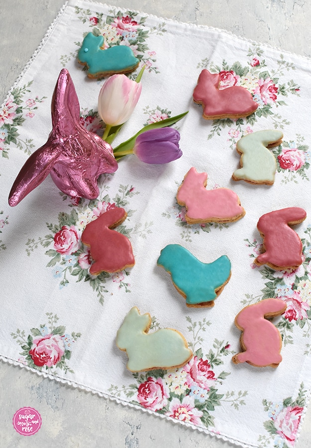 Osterhasen-Kekse in vier Pastellfarben glasiert auf Geschirrtuch mit Rosenmotiv, dazu ein Schokoosterhase in Pink und zwei Tulpenblüten