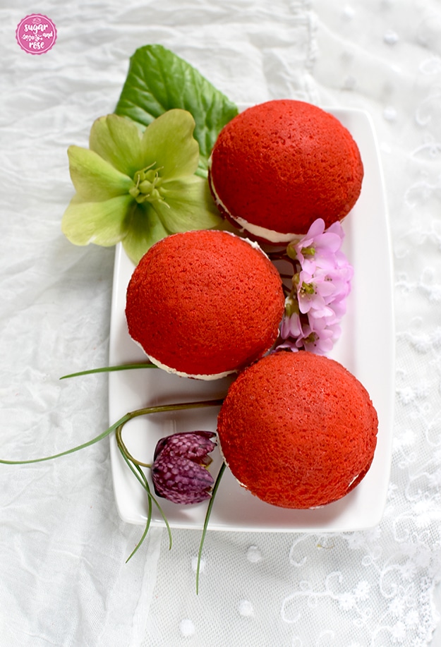 Drei Oh-Wow-Whoopie in rot-weiß-rot hintereinander auf einem weißen rechteckigen kleinen Teller aufgelegt und mit rosa Blüten und grünen Blättern dekoriert