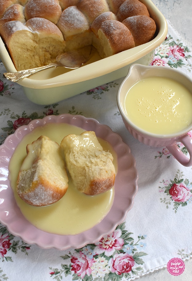 Marillenbuchteln in einer Riess-Pfanne auf Serviette mit Rosenmuster, einige Buchteln fehlen, dort liegt ein silberner Tortenheber. Davor ein rosa Keramikteller mit zwei Buchteln und Vanillesauce. Daneben eine rosa Keramikschale mit Henkel, gefüllt mit Vanillesoße