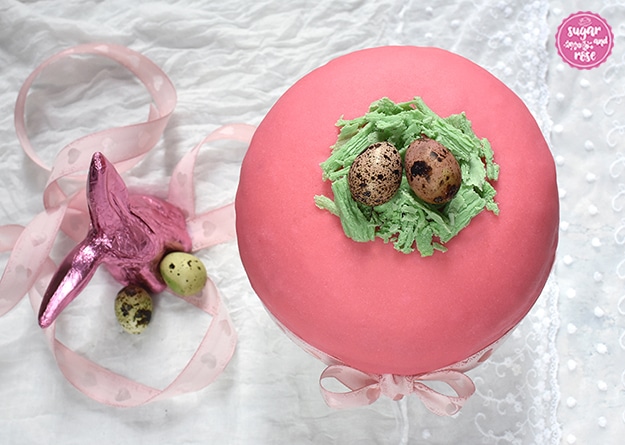 Ostertorte von oben mit grünem Schokonest und zwei bunten Wachteleiern dekoriert, daneben ein pinkfarbener Schokoosterhase in Staniol, zwei hellgrüne Wachteleier und eine hellrosa Schleife