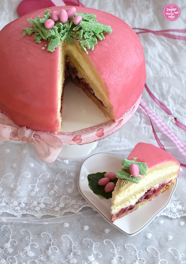 Rosa Ostertorte mit rosa und weißen Zuckereiern im Schokoosternest auf weißem Tortenständer, dekoriert mit rosa Schleife; ein Stück ist herausgeschnitten und liegt auf einem herzförmigen weißen Porzellanteller; hier sieht man die Himbeer-, Biskuit- und Vanillecremeschichten