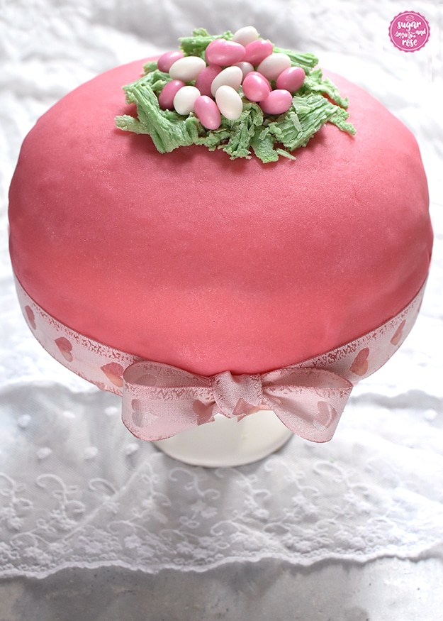 Rosa Ostertorte mit rosa und weißen Zuckereiern im Schokoosternest auf weißem Tortenständer, dekoriert mit rosa Schleife