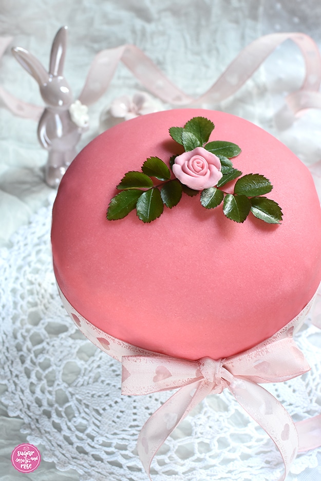 Ostertorte mit rosa Marzipanrose und grünen Rosenblättern dekoriert (Prinzessinnentorte), mit zartrosa Schleife mit Herzmotiven umwickelt, dahinter ein Porzellanosterhase