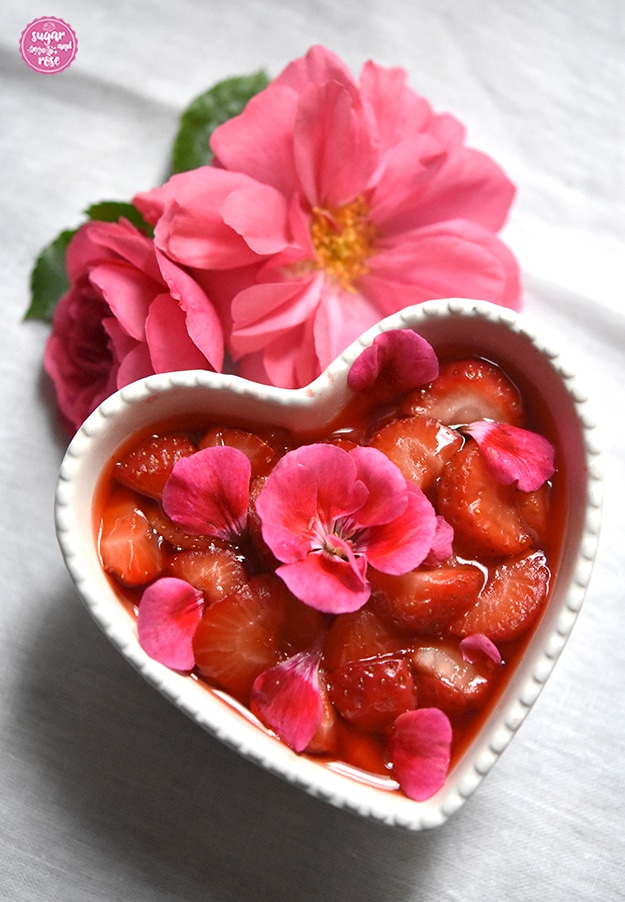 Ein herzförmige weiße Keramischale gefüllt mit Erdbeerkompott, dekoriert mit pinkfarbenen Geranienblüten, dahinter zwei pinkfarbene Rosenblüten