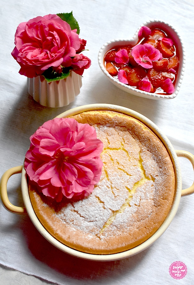 Grießauflauf in gelber Riess-Emailpfanne, dekoriert mit einer Rosenblüte, dahinter ein Erdbeer-Geranien-Kompott in herzförmiger Keramikschale, daneben eine kleine Vase mit einer pinkfarbenen Rosenblüte (Rosarium Uetersen)