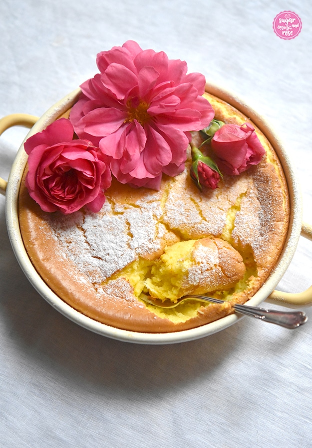 Grießauflauf in gelber Riess-Emailpfanne mit drei pinkfarbenen Rosenblüten dekoriert (Rosarium Uetersen); Auflauf mit einem silbernen Dessertlöffel an einer Stelle angestochen.