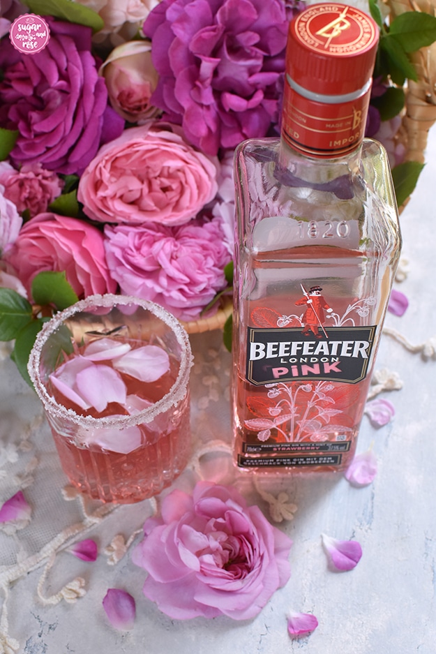 Pink Rosé Gin im Glas mit Zuckerrand, daneben eine Flasche Beefeater London Pink Gin, dahinter ein Korb mit rosa und piken Rosen