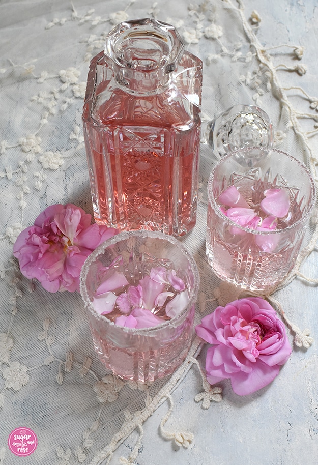 Eine geschliffene alte Karaffe mit Rosé Gin, davor zwei Gläser mit Pink Rosé-Gin und zwei Damaszenerrosenblüten