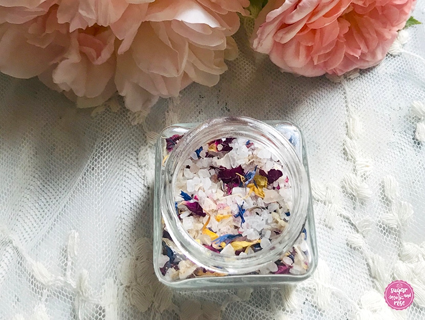 Ein kleines eckiges Glas gefüllt mit grobem Meersalz und getrockneten Blüten (Rosen, Kornblumen, Ringelblumen), dahinter einige pfirsichfarbene Rosen