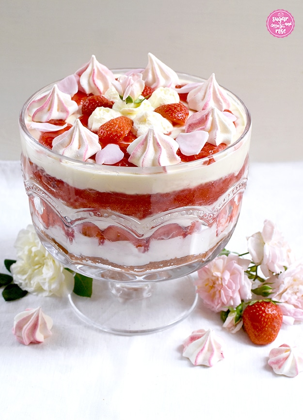 Erdbeer-Trifle in einem großen Trifle-Glas mit Fuß, rundum zartrosa und weiße Rosenblüten, rosa-weiße Baisertuffs und eine Erdbeere