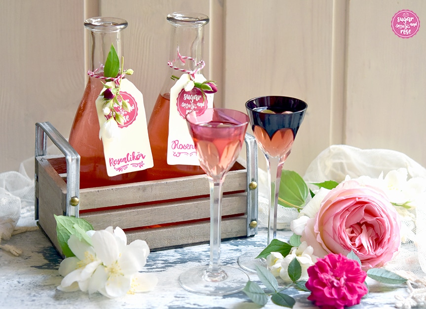 Rosenlikör in zwei Fläschchen abgefüllt und etikettiert in einer kleine hellen Holzkiste stehend, davor zwei Jugendstil-Likörgläser mit hohem Stil und Rosenblüten
