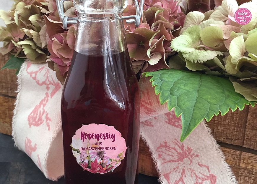 Dunkelroter Rosenessig in Flasche mit Etikett und Beschriftung "Rosenessig aus Damaszenerrosen", dahinter eine Holzbox mit Hortensien und einer breiten Stoffschleife in creme mit rosa Blütenmotiven