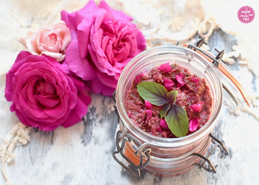 Rosenpesto in einem geöffneten Weckglas, dekoriert mit rotem Basilikum, dahinter pinkfarbene Rosenköpfe