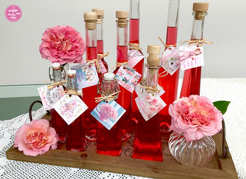 Roter Rosensirup in Flaschen abgefüllt und mit Rosenetiketten beschriftet auf einem Holztablett stehend, daneben eine bauchige kleine Glasvase mit Augusta-Luise-Rosenblütenköpfen