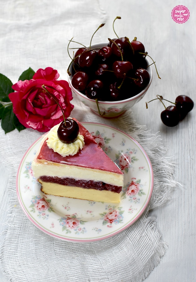 Ein Stück Topfen-Kirschtorte auf einem Desserteller mit rosa Rosenblüten und kleinen Vergissmeinnicht. Die Schichten der Torte sind gut zu sehen: unten der gelbe Topfenkuchen, dann die rote Kirschschicht, darüber die weiße Topfencreme und der zartrosa Blätterteigboden, garniert mit Schlaoberstupfen und Kirsche. Dahinter eine kleine Keramikschale mit gleichem Dekor wie der Teller, gefüllt mit Kirschen. Daneben eine pinkfarbene Henri-Matisse-Rose.