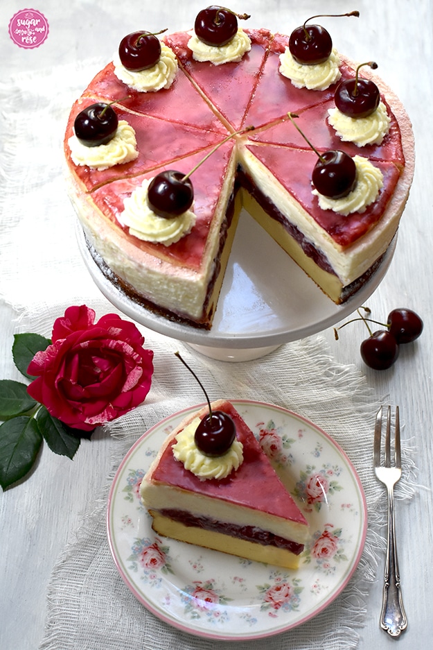 Topfen-Kirschtorte auf weißer Etagere, ein Stück ist herausgeschnitten. Vor der Torte ein Dessertteller mit kleinen Rosenmotiven, darauf ein Stück Torte, daneben Kirschen, eine Rosenblüte und eine silberne alte Dessertgabel.