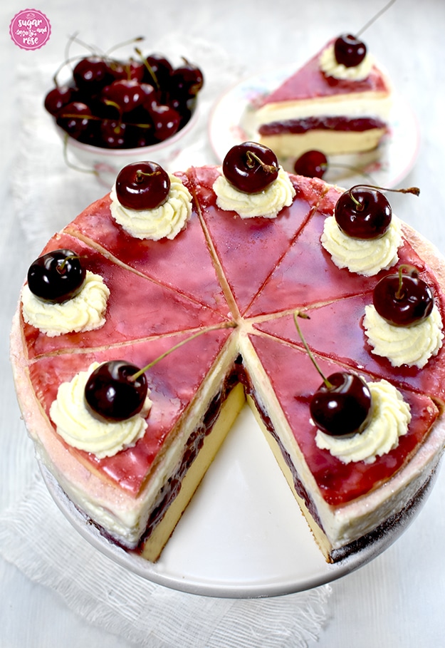 Topfen-Kirschtorte auf weißer Etagere, ein Stück ist herausgeschnitten. Dahinter eine kleine Schale mit Kirschen und ein Desserteller mit einem Tortenstück.