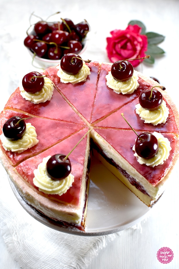 Topfen-Kirschtorte auf weißer Etagere, ein Stück ist herausgeschnitten. Dahinter eine kleine Schale mit Kirschen und eine Rosenblüte