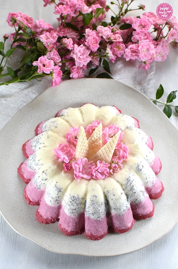 Eisgugelhupf auf unregelmäßig getöpfertem Teller, garniert mit kleinen rosa Röschen und dreieckigen Eiswaffeln, dahinter ein Zweig mit büschelweise rosa Röschen (The Fairy).