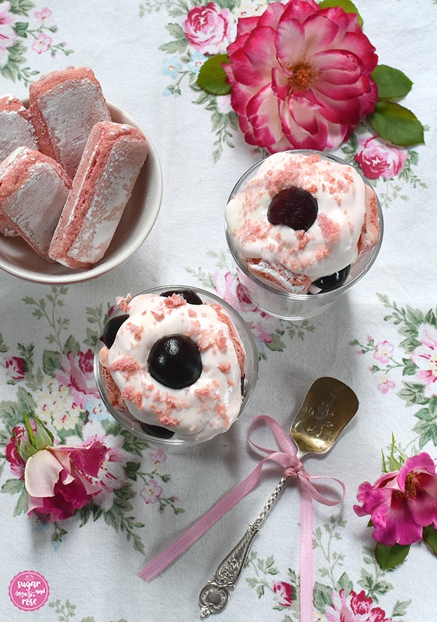 Zwei Gläser Kirschdessert, daneben eine Schale mit Biscuits Roses, davor ein silberner Dessertlöffel mit rosa Schleife, rundum rosa-weiße Rosenblüten