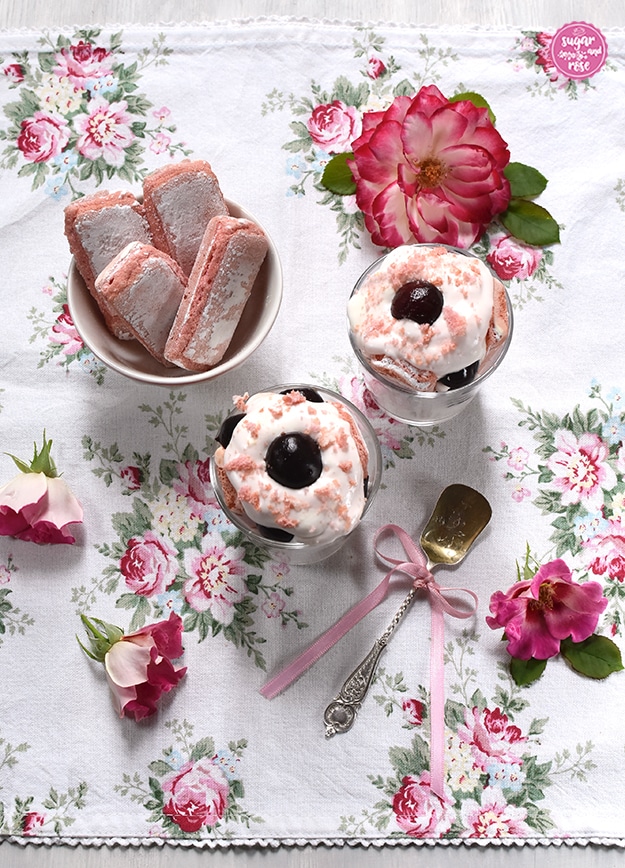 Kirschdessert im Glas, daneben eine kleine Schale mit Biscuits Roses, davor ein silberner Dessertlöffel mit rosa Schleife, rundum rosa-weiße Rosenblüten